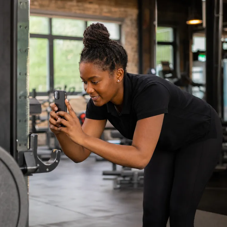 A gym staff member crouched beside a treadmill on a clean, modern gym floor, dictating a note and pointing at a detail on the machine.