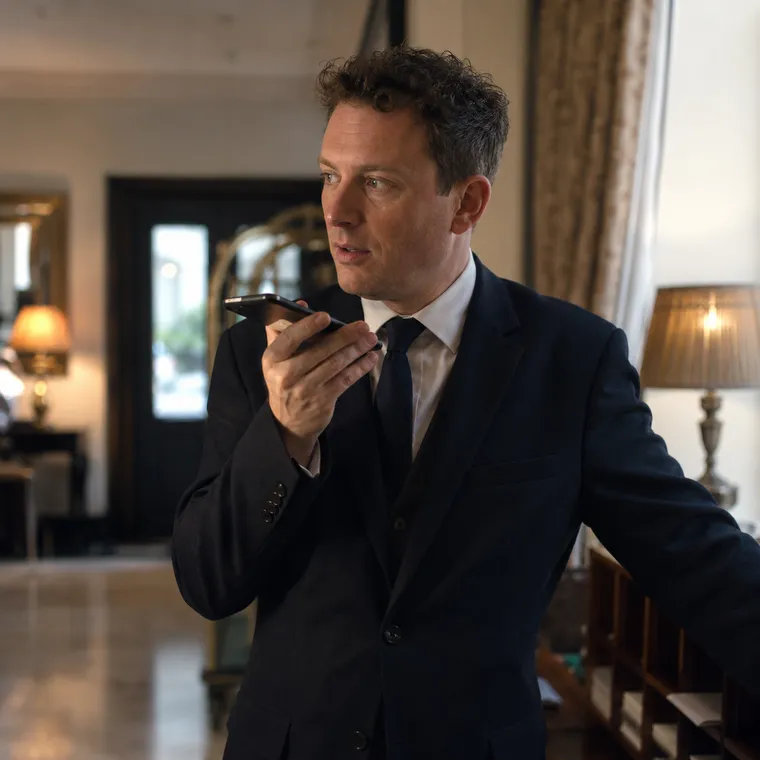 A hotel duty manager in an elegant boutique-hotel lobby, phone in hand, dictating a quick note while gesturing toward a feature wall.