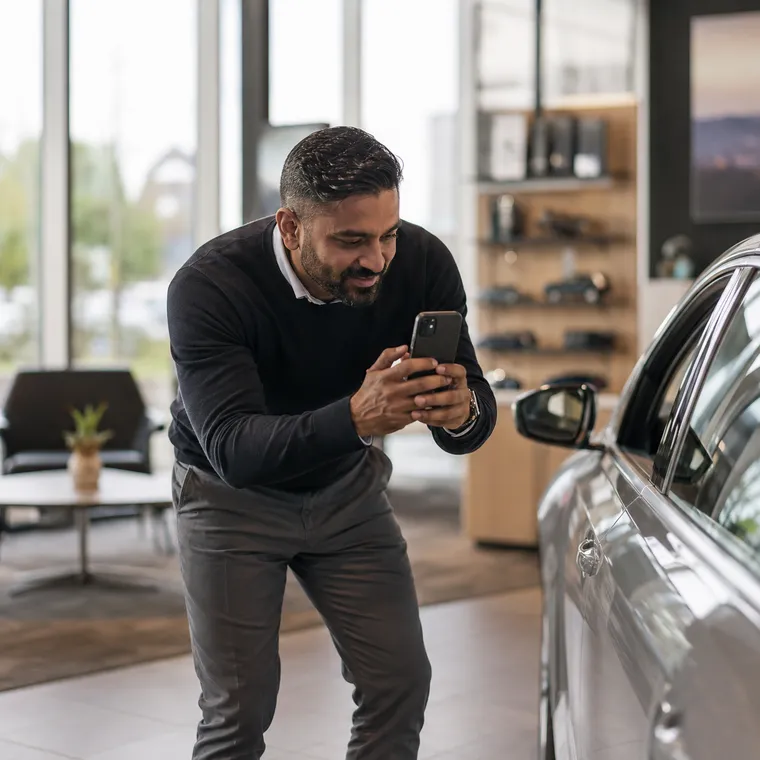 A showroom manager in a gleaming car showroom, phone in hand, dictating a note while gesturing toward a car on display.