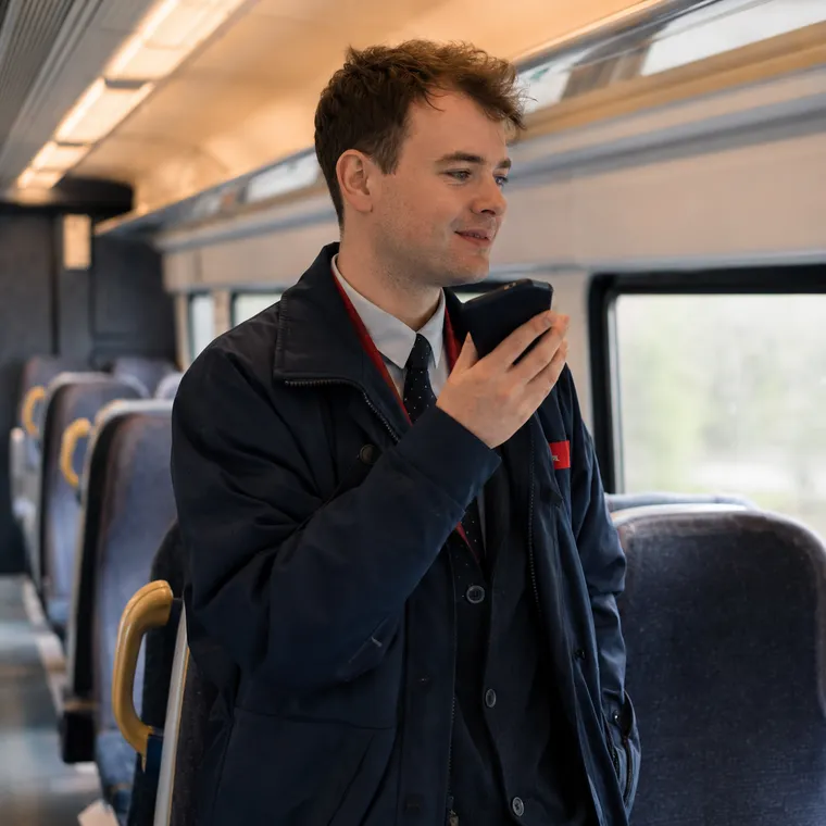 A uniformed rail staff member walking through a modern passenger carriage, phone in hand, inspecting a seat. No passengers in frame.